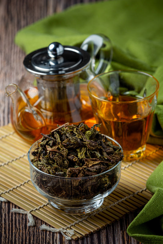 A bowl of green tea leaves with a tea pot and glass of green tea behind it, placed on a bamboo mat on a wooden surface