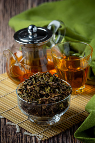 A bowl of green tea leaves with a tea pot and glass of green tea behind it, placed on a bamboo mat on a wooden surface